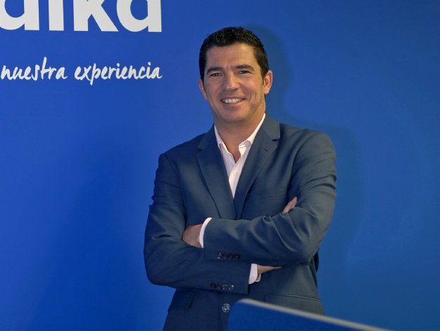 Un hombre de traje azul con brazos cruzados está de pie frente a una pared azul que muestra el logotipo de Audika y el texto ’nuestra experiencia’. El entorno sugiere una clínica auditiva.