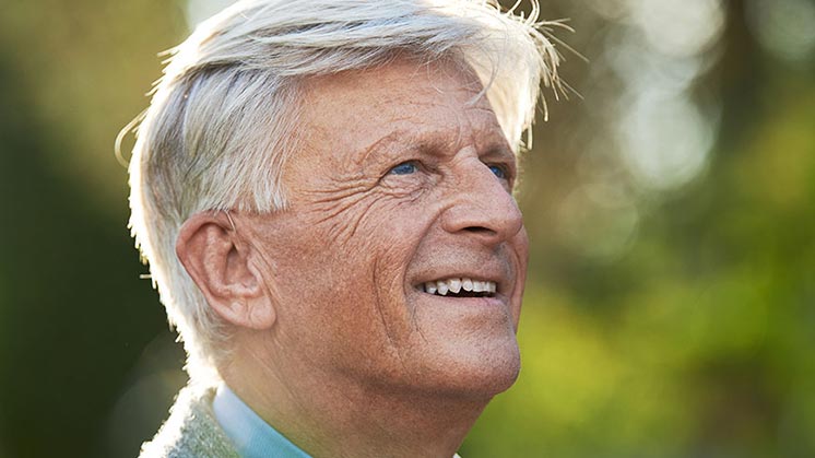 Un hombre mayor con cabello blanco muestra su oreja izquierda al descubierto, posiblemente usando un audífono discreto. El fondo es un entorno natural verde con luz suave y desenfoque.