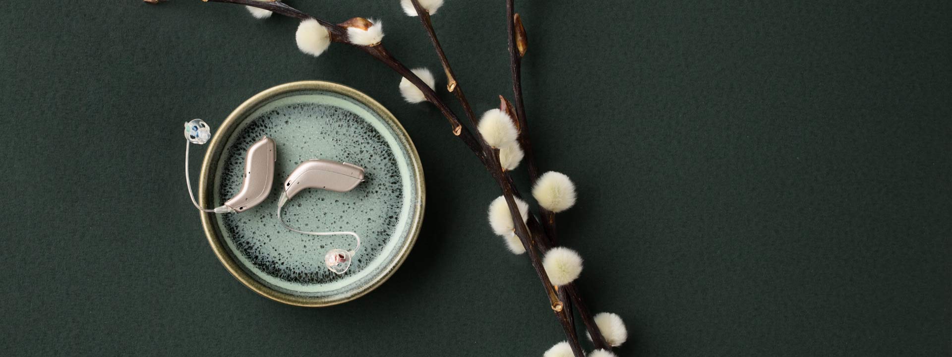 Dos audífonos modernos plateados descansan sobre un plato verde texturizado, acompañados de ramas decorativas con capullos blancos suaves, todo en un fondo verde oscuro elegante y minimalista.
