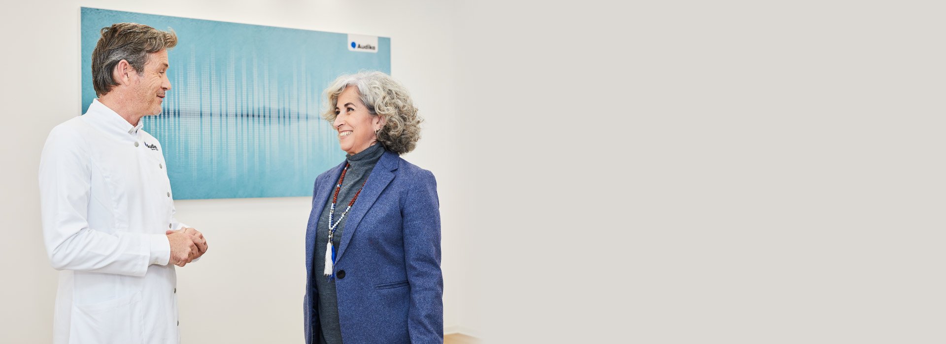 Un audioprotesista con bata blanca, identificado por el logo de Audika, conversa con una mujer mayor en una clínica de audición decorada con un cuadro abstracto azul.