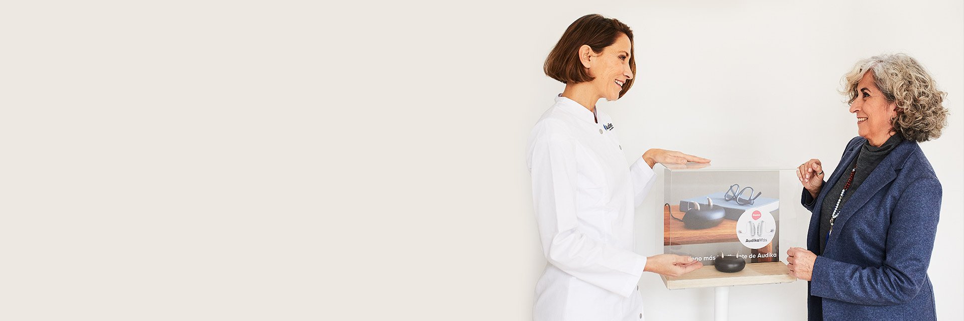 Una audioprotesista en bata blanca de Audika muestra una caja transparente con audífonos y texto ’AudikaMás’ a una mujer mayor de cabello gris, en un entorno clínico iluminado.
