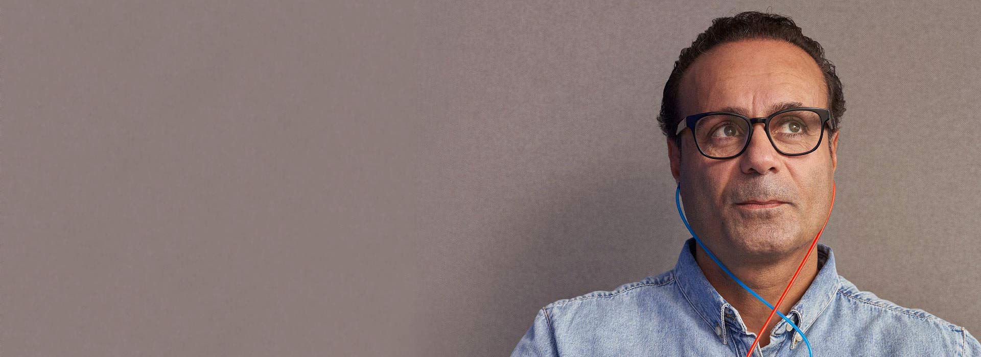 Una persona con camisa de mezclilla lleva tubos auditivos rojo y azul colgando alrededor de su cuello, en un fondo liso de color gris. No hay texto visible.