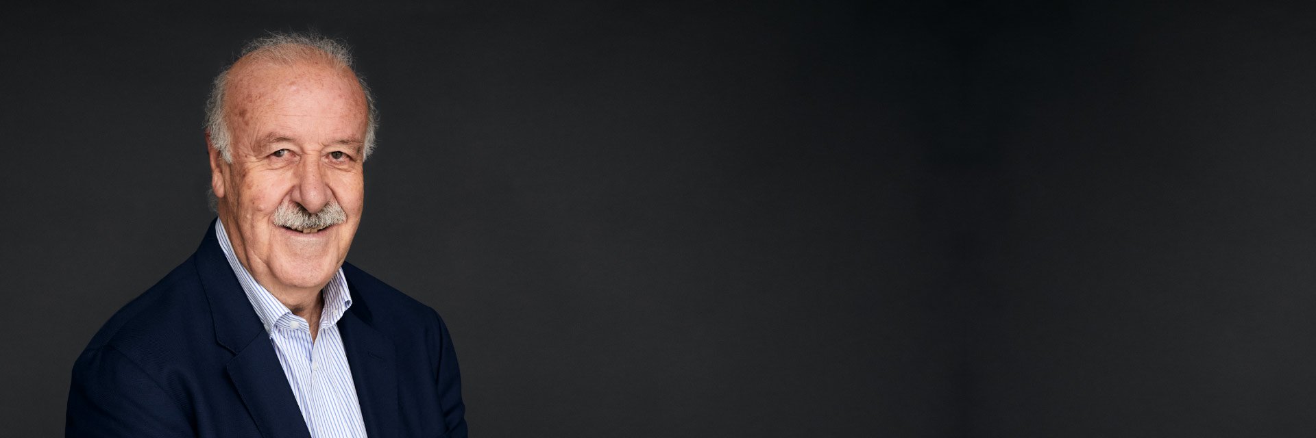Hombre mayor con cabello canoso vistiendo camisa y chaqueta oscura, posando frente a un fondo gris.