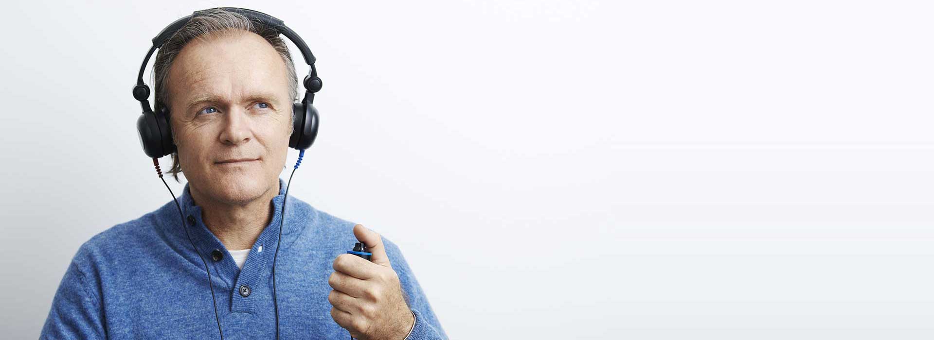 Un hombre lleva audífonos grandes y sostiene un pulsador en la mano izquierda, representando un test de audición. Fondo blanco y ropa casual azul dan un ambiente clínico.