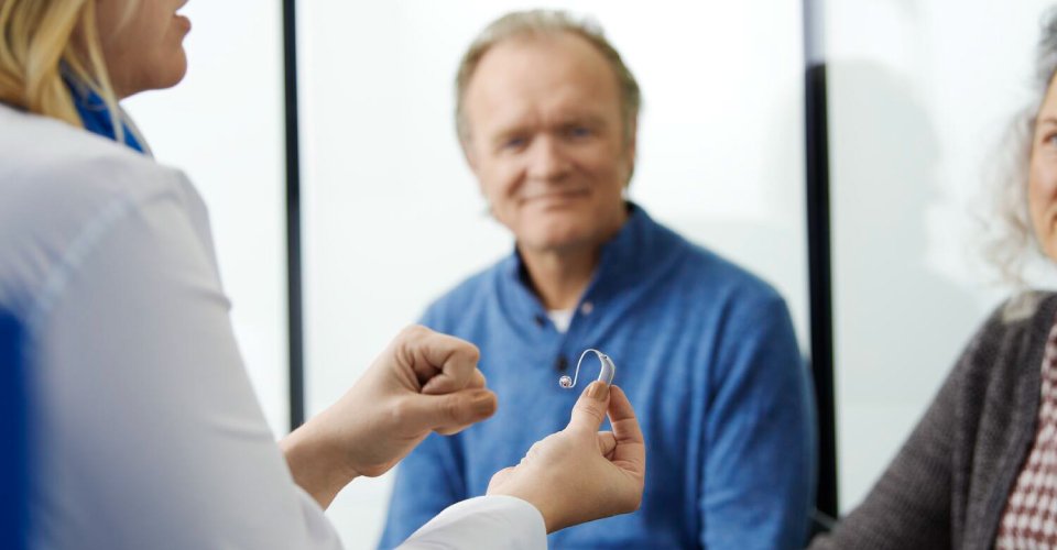 Profesional de la salud auditiva sosteniendo un audífono mientras conversa con dos pacientes adultos.