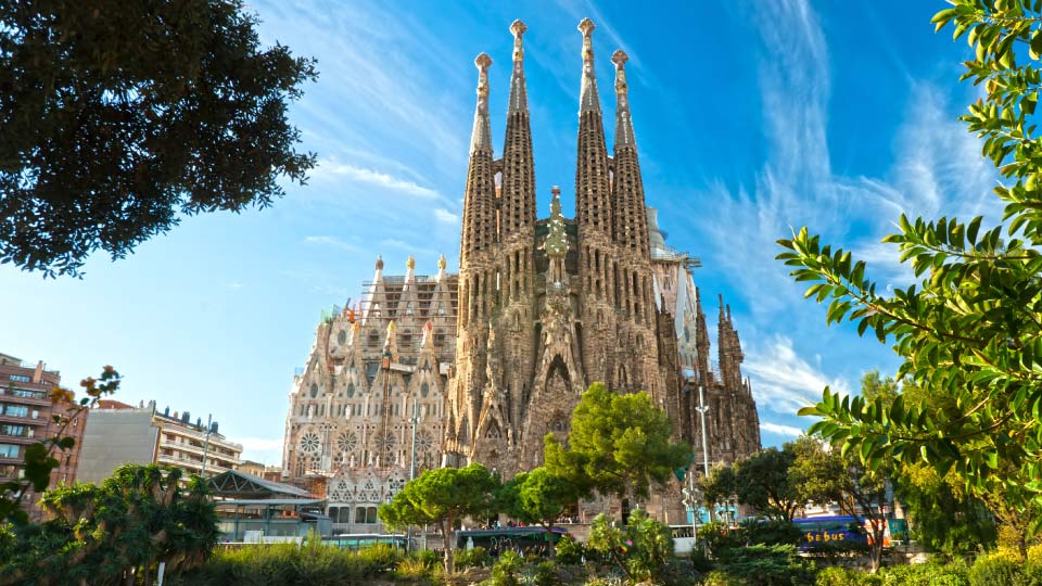 La Basílica de la Sagrada Familia, con sus icónicas torres en construcción, se alza bajo un cielo azul; rodeada de árboles, edificios urbanos y transeúntes en Barcelona.
