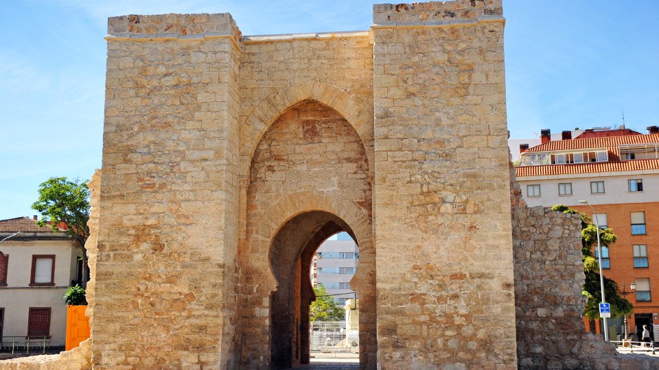 Una puerta histórica de arco de piedra, imponente y central, se alza en un entorno urbano con edificios residenciales modernos, árboles dispersos y un cielo despejado. Sin texto visible.