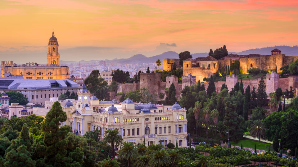 La Catedral de Málaga se eleva iluminada al atardecer mientras la Alcazaba, rodeada de vegetación, domina el paisaje urbano. Edificios históricos completan el vibrante entorno mediterráneo.
