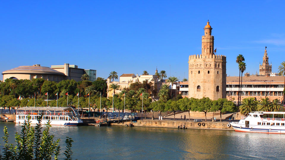 La Torre del Oro se alza junto al río Guadalquivir en Sevilla, rodeada de árboles y palmeras. Barcos turísticos navegan cerca, bajo el cielo despejado de un día soleado.