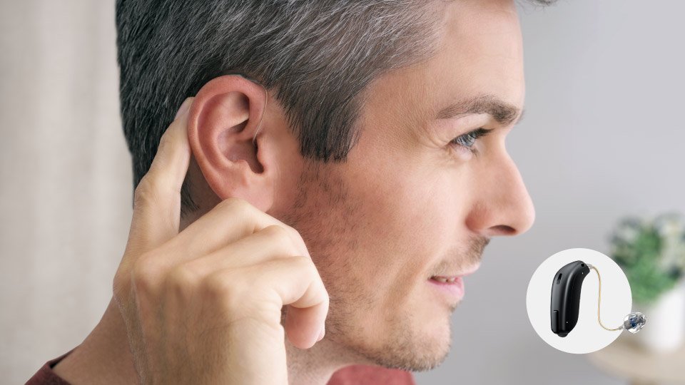 Un hombre coloca un audífono detrás de su oreja izquierda en un entorno interior luminoso. A la derecha, un recuadro muestra un audífono RIE negro en detalle.