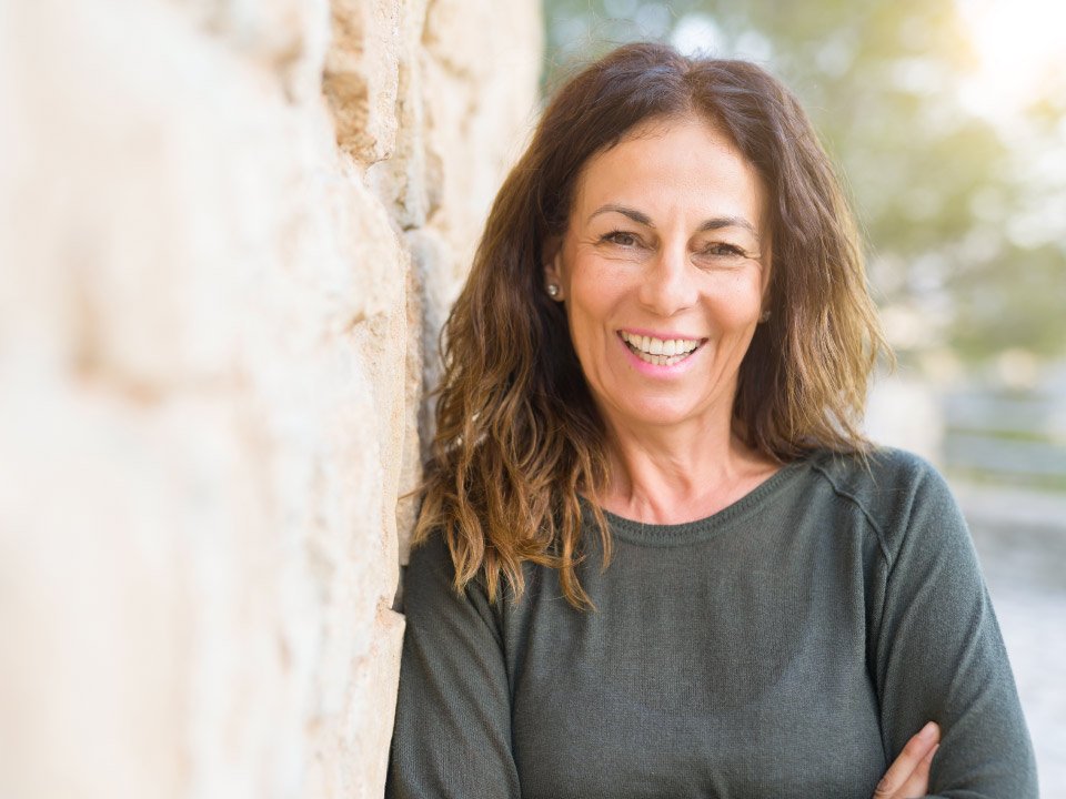 Mujer de cabello castaño, con rostro difuminado, viste un jersey verde oscuro, cruzando los brazos junto a una pared de piedra en un entorno al aire libre iluminado.