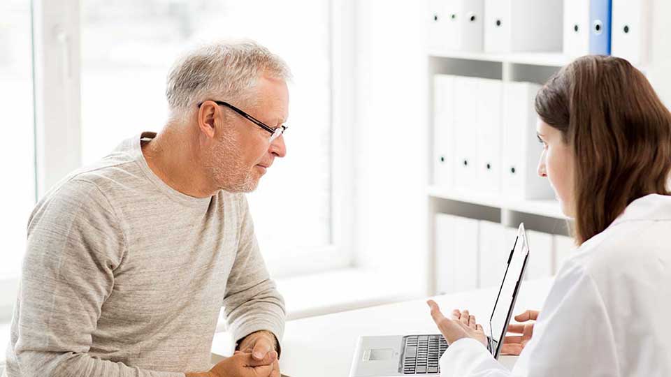 Un hombre mayor consulta con una audiológa en una clínica de audición; la profesional utiliza una computadora portátil en un ambiente iluminado y organizado con estantes llenos de carpetas.