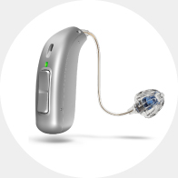 Un audífono plateado con diseño discreto de tipo RITE (receptor en el oído), con botón de control, luz LED verde y un cable fino conectado a un molde transparente con componentes internos visibles.