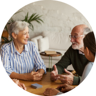 Personas mayores conversan en una mesa de madera, acompañadas de una persona más joven, en un ambiente acogedor con decoración minimalista y plantas, posiblemente en una clínica auditiva o entorno familiar.