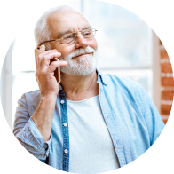 Un hombre mayor sostiene un teléfono en su mano derecha, vistiendo una camisa azul claro sobre una camiseta blanca, en un entorno iluminado con ventanas y paredes de ladrillo rojo.