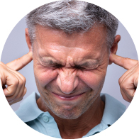 Un hombre de cabello canoso, con su rostro difuminado, se tapa ambos oídos usando sus dedos índices. Fondo liso color gris.