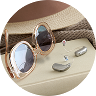 Un par de audífonos discretos y modernos se colocan sobre una superficie beige junto a gafas de sol grandes y un sombrero, en un contexto relajado y casual.