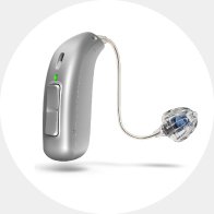 Un audífono plateado con diseño discreto de tipo RITE (receptor en el oído), con botón de control, luz LED verde y un cable fino conectado a un molde transparente con componentes internos visibles.