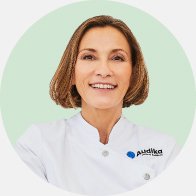 Una audioprotesista con bata blanca aparece en un fondo verde claro; el uniforme muestra el logotipo de Audika, resaltando su especialización en audífonos y cuidado auditivo profesional.