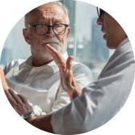 Dos personas conversan en un entorno iluminado con ventanas grandes y vista urbana. Una gesticula mientras habla, sugiriendo una consulta, posiblemente en un contexto de audición o asesoramiento en audífonos.