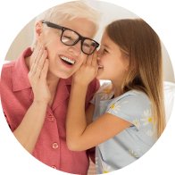 Una mujer mayor en camisa rosa sostiene su oreja mientras una niña con vestido azul claro le susurra al oído en un entorno doméstico iluminado. No hay texto visible.