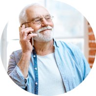 Un hombre mayor sostiene un teléfono en su mano derecha, vistiendo una camisa azul claro sobre una camiseta blanca, en un entorno iluminado con ventanas y paredes de ladrillo rojo.