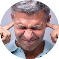 Un hombre de cabello canoso, con su rostro difuminado, se tapa ambos oídos usando sus dedos índices. Fondo liso color gris.