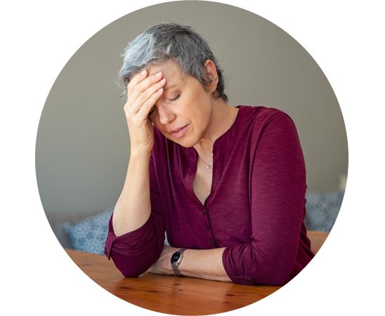 Mujer mayor con cabello canoso inclinada hacia adelante mientras se sujeta la cabeza, mostrando expresión de malestar o dolor.