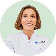 Una audioprotesista con bata blanca y logo de Audika en el pecho, de pie sobre un fondo verde claro. No se distinguen detalles del rostro debido al desenfoque.