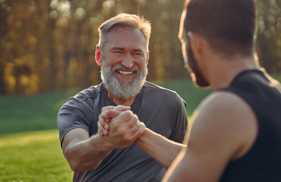 Dos hombres estrechan sus manos firmemente en un gesto de camaradería o apoyo. Ambos llevan ropa deportiva; el fondo muestra un parque verde con árboles y luz solar suave.