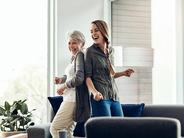 Dos mujeres, una mayor y otra joven, bailan alegremente en una sala iluminada y moderna con sofás, cojines azules y plantas decorativas cerca de una ventana grande.
