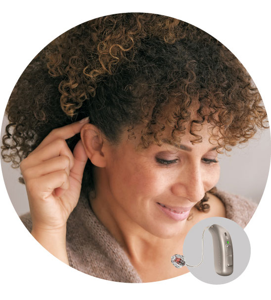 Un audífono detrás del oído se coloca discretamente por una mano femenina; el contexto sugiere un entorno doméstico. En la esquina, un detalle amplía el diseño del dispositivo auditivo.