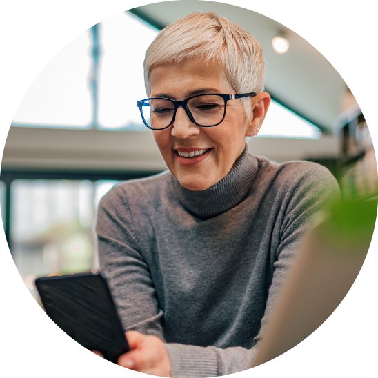 Una persona de cabello canoso y gafas sostiene un teléfono móvil en un espacio moderno y bien iluminado, posiblemente una clínica o cafetería. No hay texto visible ni audífonos identificables.