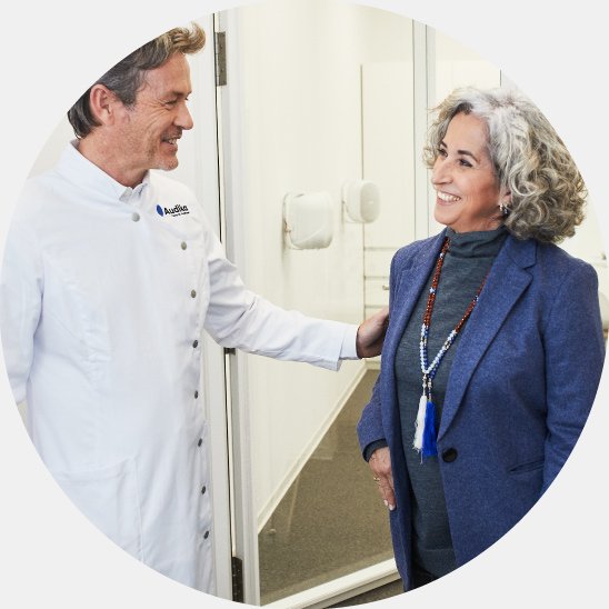 Un audioprotesista con bata blanca y logotipo de Audika conversa con una mujer mayor en un audífono azul, en el interior de una clínica auditiva moderna con paredes blancas.