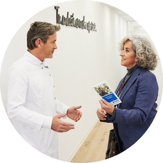 Un audioprotesista con bata blanca de Audika conversa con una mujer mayor de cabello gris que sostiene un folleto titulado ’Vuelta al sonido natural’ en un pasillo iluminado. En la pared se lee el texto: ’Tu audición en buenas manos’.