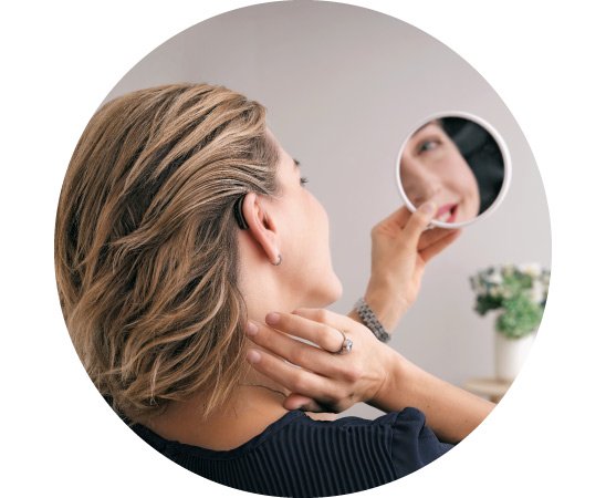 Una mujer con cabello rubio corto utiliza un espejo de mano, revisando detrás de su oreja donde lleva un audífono discreto. Fondo difuminado con una planta decorativa en una clínica auditiva.