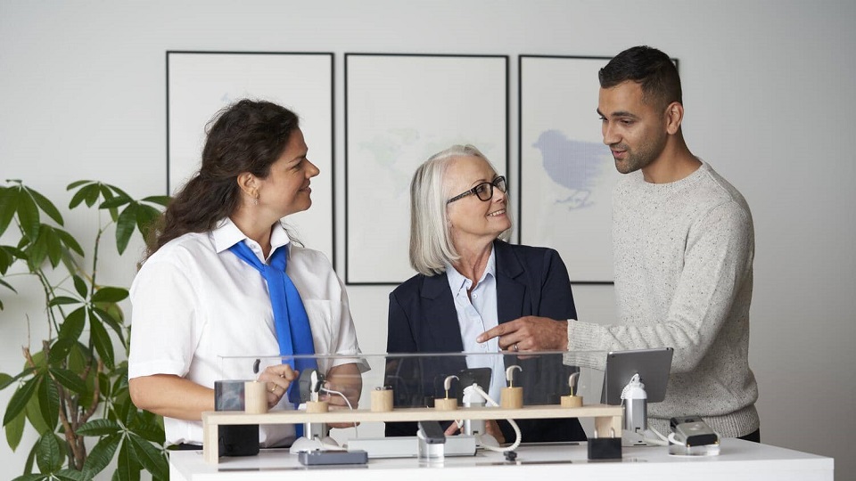 Une audioprothésiste explique les options d'appareils auditifs à une patiente et à son fils dans un centre auditif.