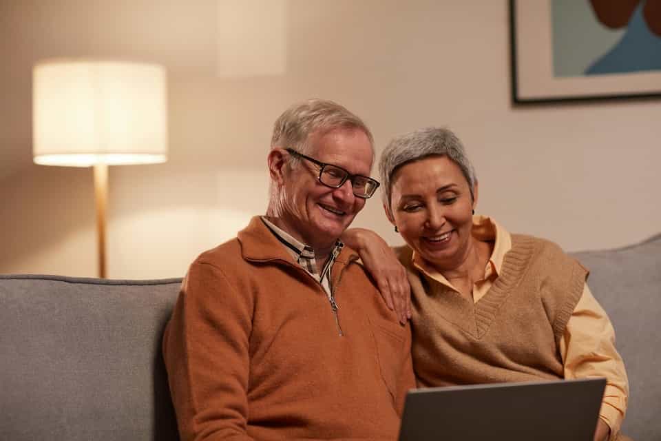 Un couple de seniors assis sur un canapé utilise une tablette dans un espace de vie chaleureux.