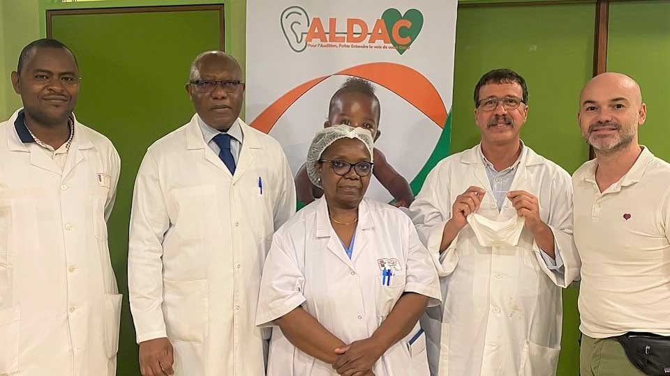 Des professionnels Audika participant à une mission humanitaire au Cameroun devant une affiche de l’association ALDAC.