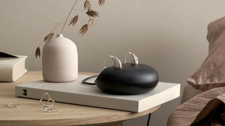 Deux appareils auditifs rechargeables en train de charger sur un socle noir, posés sur un livre dans un décor intérieur épuré.