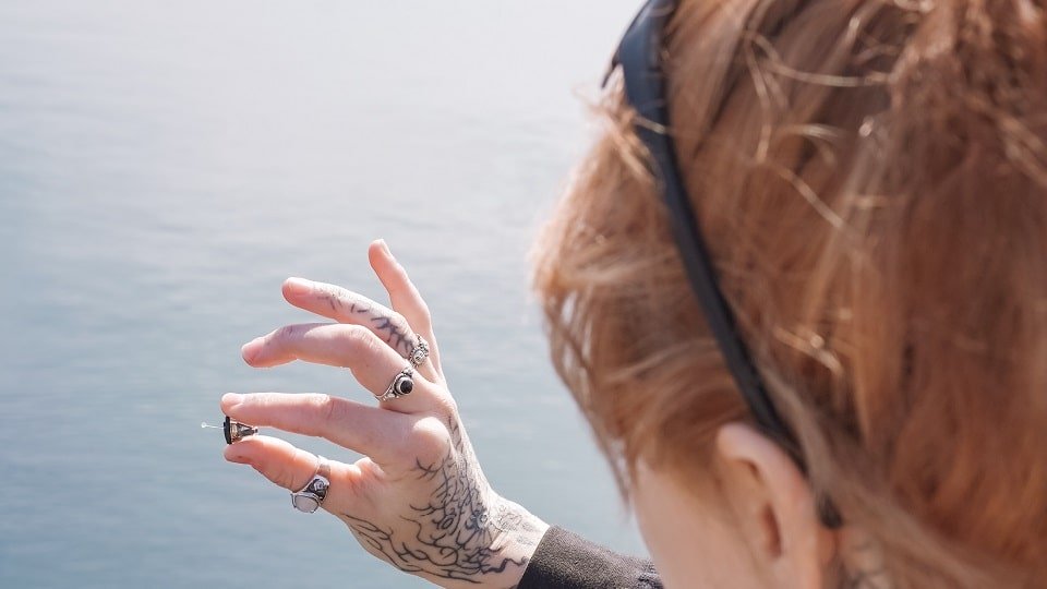Une femme aux cheveux roux et tatouages tient des appareils auditifs intra-auriculaires devant un paysage marin.