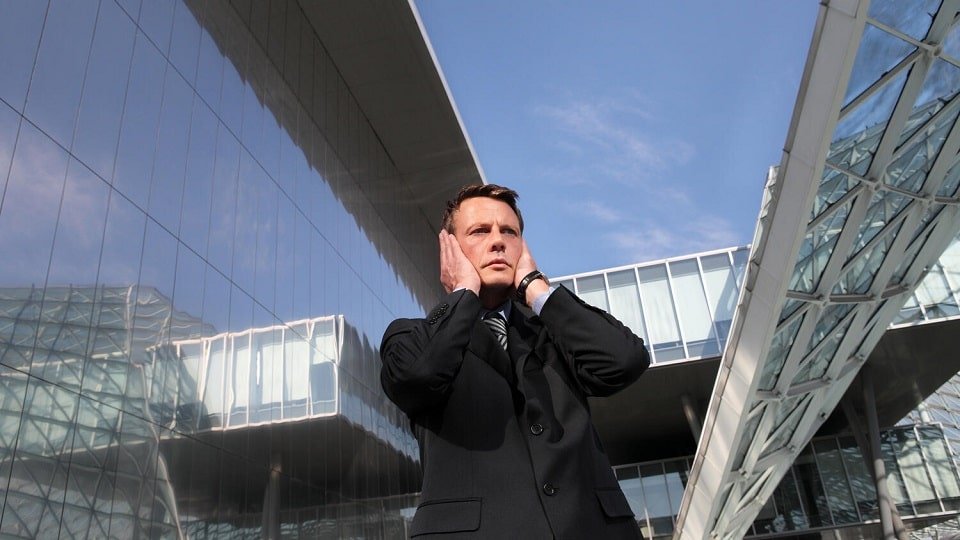 Une personne en costume tenant sa tête avec ses mains devant un bâtiment moderne en verre.
