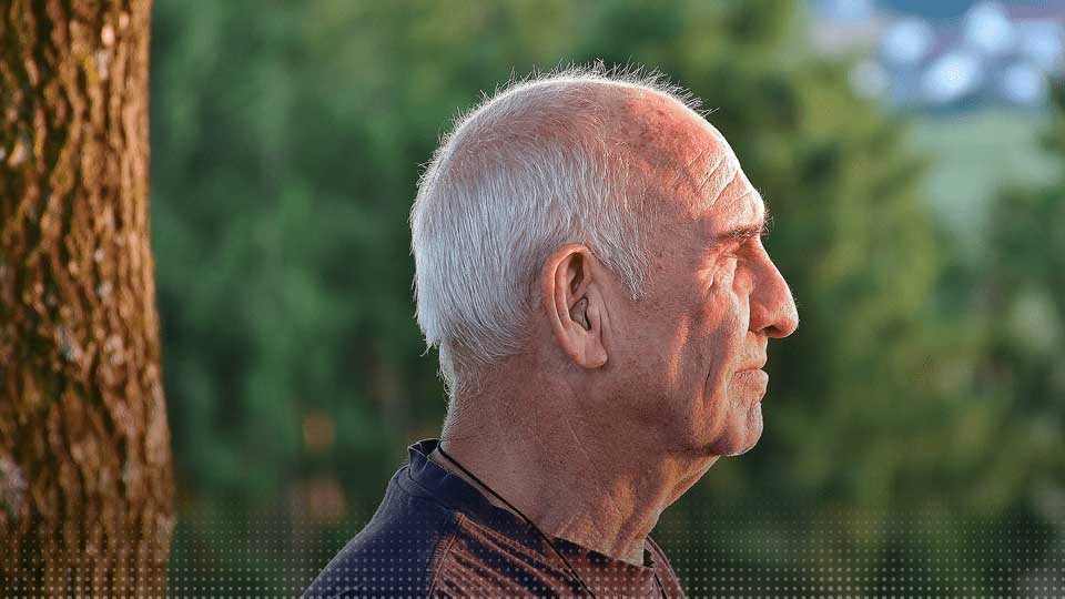 Un homme âgé portant un appareil auditif, vu de profil, en extérieur.