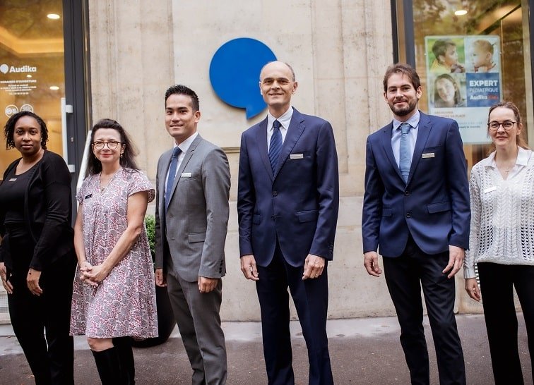 L’équipe professionnelle devant un centre auditif Audika, portant des tenues élégantes et se tenant devant le logo bleu de l’entreprise.