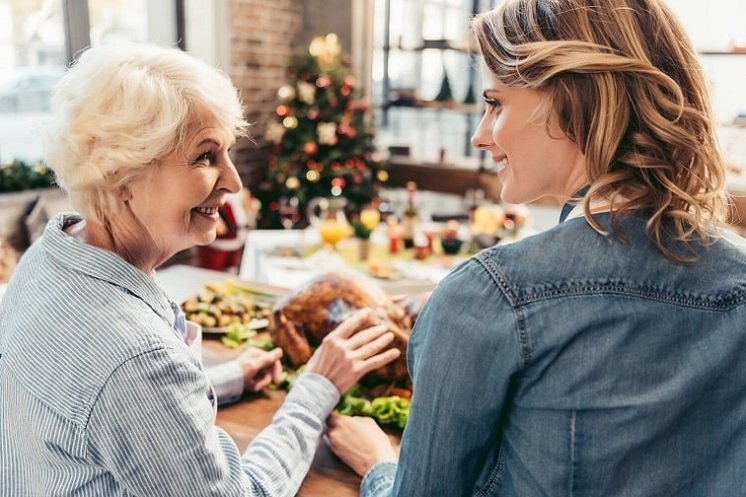 Une personne âgée portant un appareil auditif échange avec une jeune femme lors d’un repas de fête devant un sapin de Noël décoré.