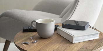 Des appareils auditifs mini-contours sur une table basse en bois avec un livre, une tasse grise et un appareil électronique dans un salon lumineux.