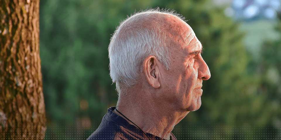 Un homme senior debout à l'extérieur, de profil, avec des arbres en arrière-plan.