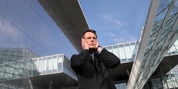 Un homme en costume tenant ses oreilles devant un bâtiment moderne, exprimant une gêne due au bruit ou aux acouphènes.