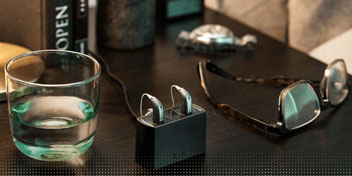 Deux aides auditives rechargeables posées sur leur station d’accueil, à côté d’un verre d’eau et de lunettes sur une table.