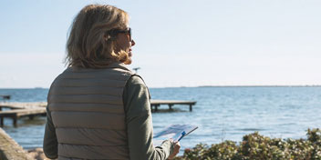 Femme avec un gilet matelassé tenant un carnet face à un lac avec des pontons.